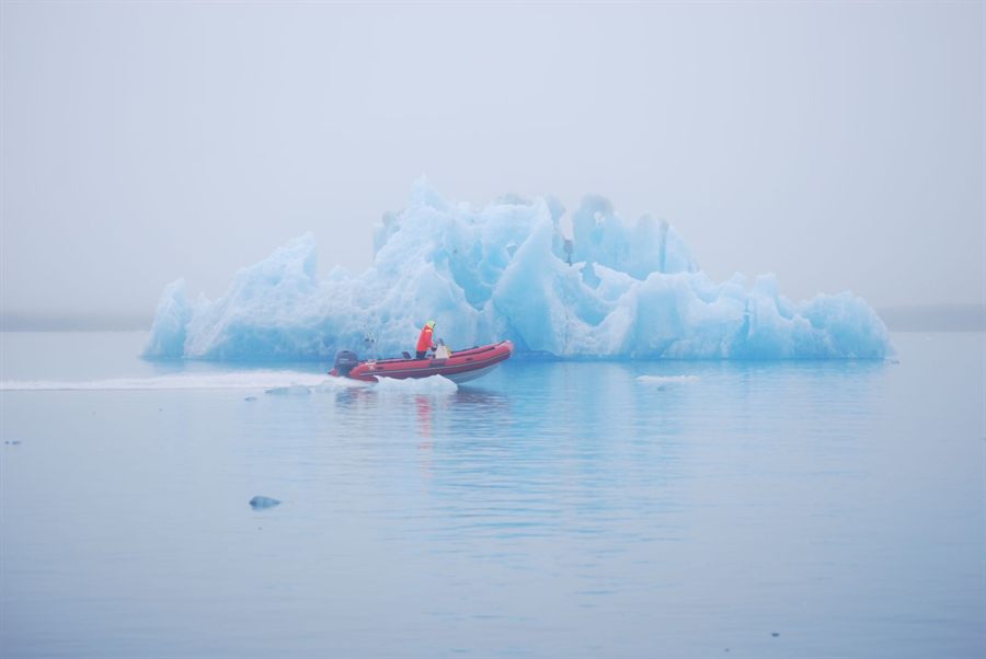 Small red boat beside a large blue iceberg in calm water