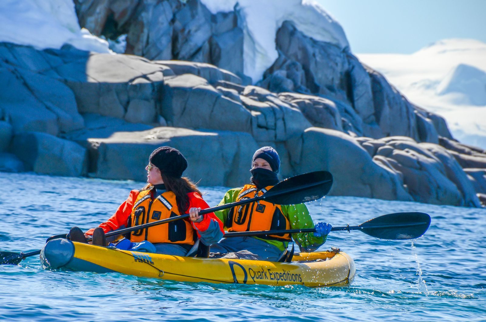 Two kayakers in polar gear paddling below an icy cliff on an expedition voyage