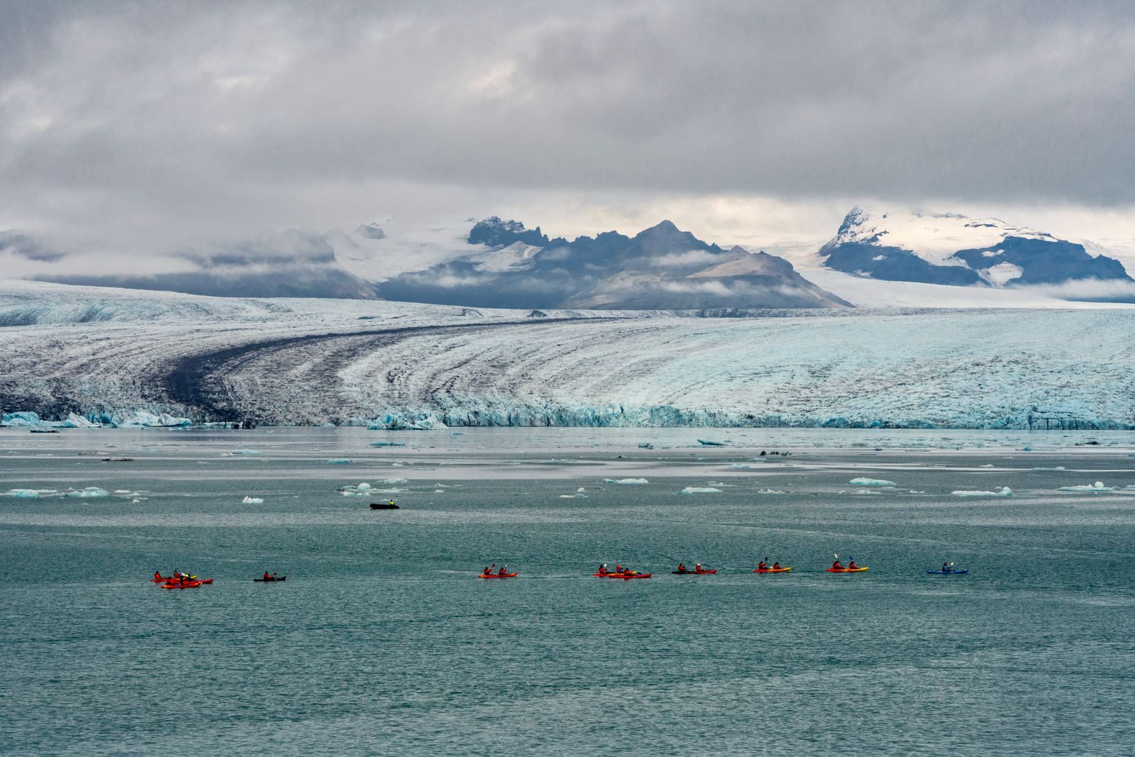 Line of kayaks crossing a bay below a wide glacier